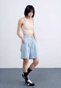 Top court beige, shorts en denim plissés bleu clair, et baskets noires avec accents blancs. Le mannequin se tient les mains dans les poches sur un sol gris.