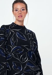 Blusa nera con motivo a foglia bianca e blu, collo alto e maniche strutturate, realizzata in un tessuto leggero con una consistenza liscia.