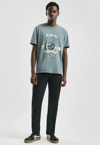 Młody mężczyzna w wyblakłej niebieskiej koszulce z grafiką, czarnych dżinsach i czarnych trampkach z białymi akcentami, stojący na tle gładkiego białego tła.