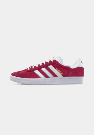 Rode suede sneaker met witte leren drie stripes en logo. Heeft een rubberen midsole met een gestructureerd oppervlak en een afgeronde neus.