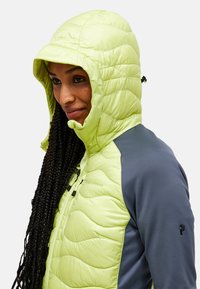 Lys grøn puffy jakke med hætte, der har et quilted design, lynlåslommer og grå sidepaneler. Glat tekstur, justerbar hætte.