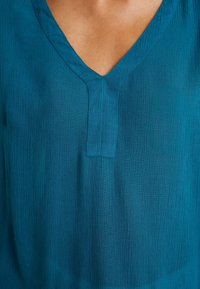 Blouse bleu sarcelle à col en V en tissu texturé léger ; motif vertical subtil ; coupe décontractée avec détail de patte à l'encolure.