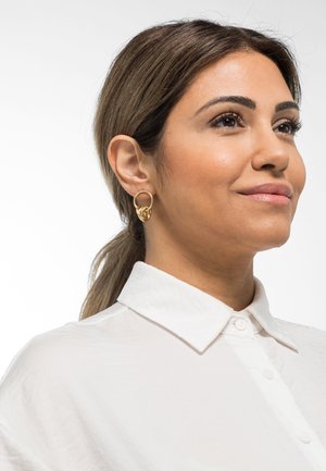 Orecchino a cerchio d'oro con un design arricciato. Camicia bianca con colletto, texture liscia e bottoni visibili. Soggetto rivolto leggermente verso destra.
