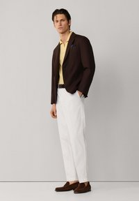 Uomo che indossa un blazer marrone scuro, camicia gialla, pantaloni bianchi e mocassini marroni, in piedi davanti a uno sfondo grigio chiaro.