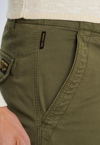 Oliveno zelene vojaške hlače s ojačenimi šivi, stranskim žepom in tkaninsko etiketo. Material se zdi trpežen z gladko teksturo.