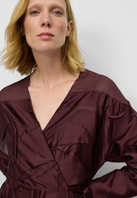 Blond kvinna i en djup burgundy satin klänning med V-halsringning, poserande mot en enkel ljusgrå bakgrund.