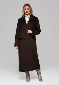 Nainen, jolla on pitkät vaaleat hiukset, pukeutuneena tummanruskeaan tuplarivistä takkiin, valkoiseen rusetti-paitaan ja käärmeennahkakuosiset polven yli ylettyviin saappaisiin harmaata taustaa vasten.