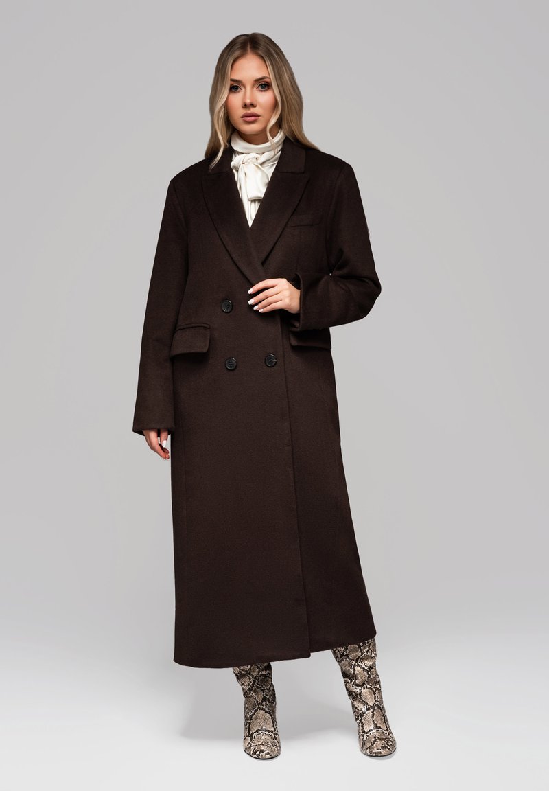 Nainen, jolla on pitkät vaaleat hiukset, pukeutuneena tummanruskeaan tuplarivistä takkiin, valkoiseen rusetti-paitaan ja käärmeennahkakuosiset polven yli ylettyviin saappaisiin harmaata taustaa vasten.