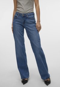 Jeans de pierna ancha de mezclilla azul, con bolsillos frontales, cierre de botón y cintura de altura media, combinados con zapatos negros de punta.