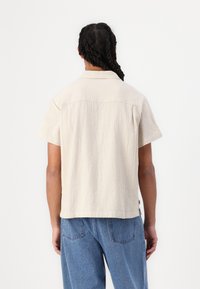 Short-sleeve beige shirt with a textured fabric. Features a collar and a straight hem. Worn with blue denim pants.