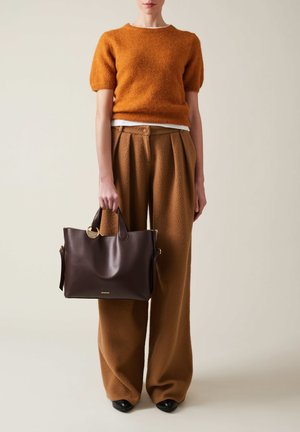 Sac en cuir marron avec des poignées rondes, un accent en métal doré, et une finition lisse, porté par une personne vêtue d'un pull et d'un pantalon couleur rouille.