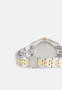 Silver- och guldtonad metallklocka med rund klocka, texturerad armband och en klassisk spänne. Baksidan är slät utan synliga märkningar.