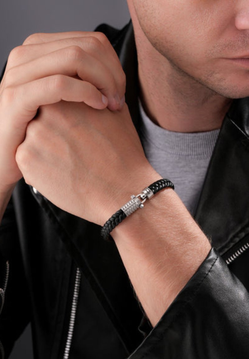 Bracciale in pelle nera intrecciata con una chiusura in metallo argentato caratterizzata da rifiniture testurizzate, indossato su un polso contro uno sfondo scuro.