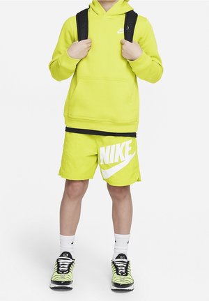 Dziecko ubrane w neonowo żółtą bluzę z kapturem i spodenki Nike z białym logo Nike, białe skarpetki, czarno-żółte buty sportowe, trzymając czarne szelki plecaka.