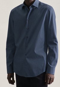 Camisa de botones azul con un estampado, de cuello puntiagudo, que presenta un sutil diseño geométrico y mangas largas. Los botones son oscuros y están en la parte frontal.