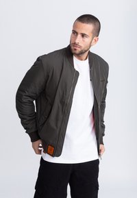 Kurtka bomberka w ciemnozielonym kolorze, wykonana z gładkiego nylonu, z prążkowanym kołnierzem, zamkiem błyskawicznym, bocznymi kieszeniami i pomarańczowym akcentem na etykiecie.