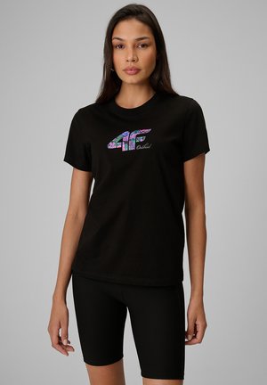 Kobieta z długimi ciemnymi włosami, ubrana w czarną koszulkę z kolorowym logo "4F Orchid" oraz w czarne, prążkowane kolarskie szorty, stoi na tle szarego tła.