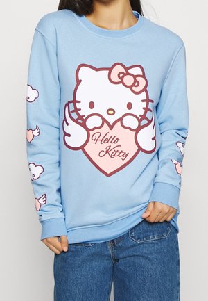 Persoon draagt een lichtblauwe trui met een grote Hello Kitty die een hart vasthoudt met tekst en gevleugelde harten op de mouwen.