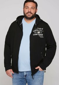 Chaqueta negra con capucha y texto gráfico en blanco y elementos de diseño. Cuenta con un cierre de cremallera y cordones ajustables. Llevada sobre una camisa de color azul claro.