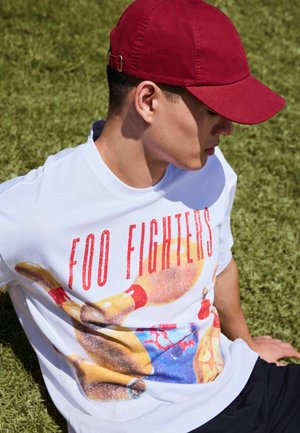 Jaunas vyras dėvi baltą Foo Fighters grupės marškinėlį ir raudoną beisbolo kepuraitę, sėdi ant žolės ir žiūri žemyn į šoną.