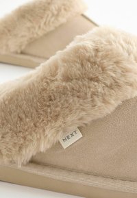Pantofole beige con una parte superiore in pelliccia morbida e un corpo in suede morbido. Presentano un piccolo cartellino "NEXT" sul lato e una suola in gomma texturizzata.