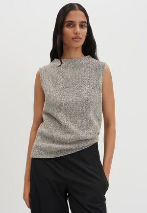 Femme aux longs cheveux foncés portant un haut gris texturé sans manches et un pantalon noir sur un fond clair uni.