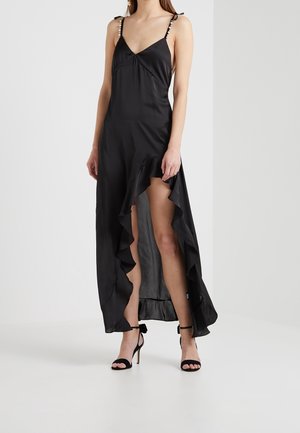 Kobieta ubrana w czarną, satynową suknię maxi z falbanami i wysokim rozcięciem, zawiązywane na ramionach ramiączka oraz czarne sandały na wysokim obcasie z paskami.