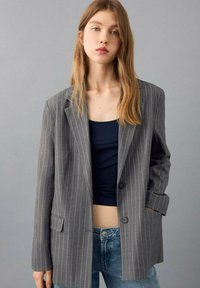 Grijze blazer met krijtstreep en een relaxte pasvorm, single knoopsluiting en twee voorzakken, gedragen over een marineblauw crop top en lichtblauwe jeans.
