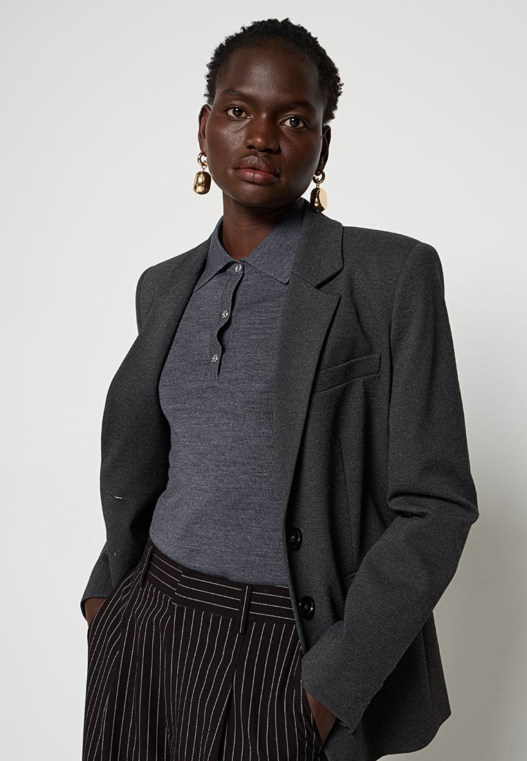 Blazer grigio elegante sopra un polo grigio scuro, caratterizzato da un colletto e dettagli con bottoni. Pantaloni a vita alta a righe completano l'outfit.