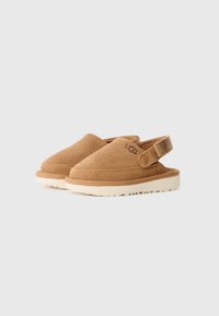 Chaussures slip-on en cuir suédé marron avec une semelle extérieure blanche, un bout arrondi et une sangle réglable au niveau du talon. Présente des accents de marque.