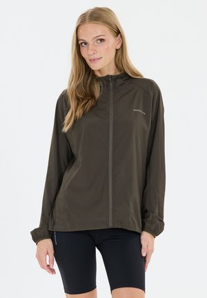 Nuori nainen, jolla on päällään tummanruskea vetoketjullinen kevyt takki ja mustat pyöräilyshortsit, seisomassa yksinkertaisen valkoisen taustan edessä.