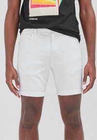 Pantalones cortos de denim blanco con puños enrollados, diseño de cinco bolsillos y una textura suave. Llevados con una camiseta gráfica negra que presenta un estampado colorido.