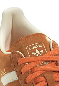 Nærbillede af orange Adidas Gazelle sneakers med orange snørrebånd, ruskindsoverflade og hvid indvendig foring.