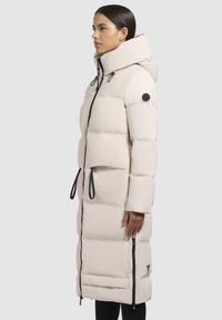 Woman wearing a long, pale beige puffer coat with a high collar, black zipper, side zipper details, and drawstring accents.