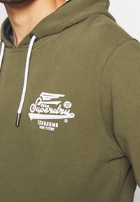 Olivově zelená mikina s kapucí s bílým logem a nápisem. Má kapuci se šňůrkou a měkký povrch. Obsahuje stříbrné kovové doplňky.