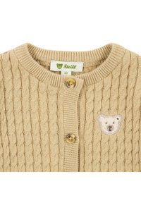 Bežový pletený kardigan s texturovaným vzorem, kulatým výstřihem, dvěma zlatými knoflíky a krémovou nášivkou medvídka na levé straně.