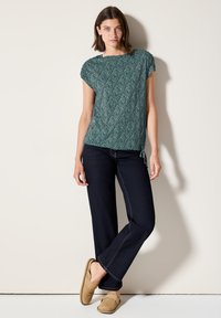 Haut à manches courtes à motifs verts avec un col arrondi, associé à un jean évasé bleu foncé et des chaussures à enfiler marron clair.