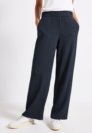 Marineblau gefaltete weite Hosen mit einem elastischen Bund und zwei Seitentaschen, kombiniert mit weißen Sneakers vor einem neutralen Hintergrund.