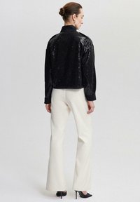 Jachetă bomber cu paiete negre, cu guler, umeri rotunzi și deschidere frontală, purtată peste pantaloni cremoși cu evazare și tocuri negre.