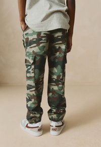 Camouflage-Cargohosen in Grün, Braun und Beige mit elastischen Bündchen, seitlichen Taschen und weiter Passform, kombiniert mit weißen und grauen Sneakers.