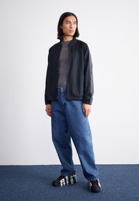 Sort zip-up jakke med opretstående krave, grå bodysuit, løse blå jeans og sorte sneakers med sølvdetaljer, imod en hvid baggrund.
