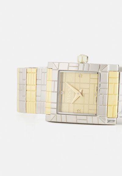 Ruudukujuline randme kell, millel on kuld- ja hõbedane geomeetriline metallrihm, tekstureeritud äär ja helekuldne ketas, millel on kolm väikest teemanti.