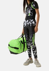 Sportoutfit med en topp i svart och vitt mönster, svarta leggings med gröna detaljer och en ljusgrön duffelväska.