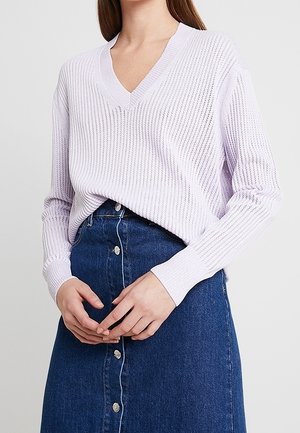 Frau trägt einen hellvioletten V-Ausschnitt-Strickpullover, der in einen dunkelblauen Jeansrock mit Knopfleiste gesteckt ist, Hände vor der Taille verschränkt.