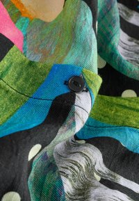 Barevná látka s kombinací modré, zelené, růžové a černé. Obsahuje puntíky, točivé vzory a detail s černým knoflíkem.