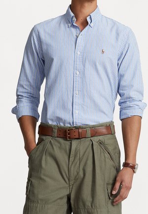 Mężczyzna ubrany w jasnoniebieską koszulę w paski z podwiniętymi rękawami, oliwkowozielone bojówki, brązowy pasek oraz skórzany zegarek na nadgarstku.