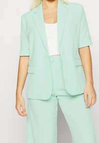 Mintgroene op maat gemaakte blazer met korte mouwen; voorzien van enkele knoopsluiting en notch revers. Gecombineerd met bijpassende mintkleurige broek.
