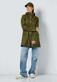 Olivno zelena vodoodporna jakna s kapuco, pritisnimi gumbi in sprednjimi žepi, v kombinaciji s svetlimi modrimi kavbojkami in vključeno pokrivalo.