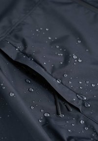 Vattentålig svart jacka i tyg med en dragkedjeförsedd ficka, där vattendroppar fastnar på den släta, matta ytan.