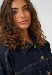 Frau mit lockigem braunem Haar und grünen Augen, die eine dunkelblaue Jeansjacke mit bronzenen Knöpfen trägt und leicht lächelt, vor einem hellen Hintergrund.